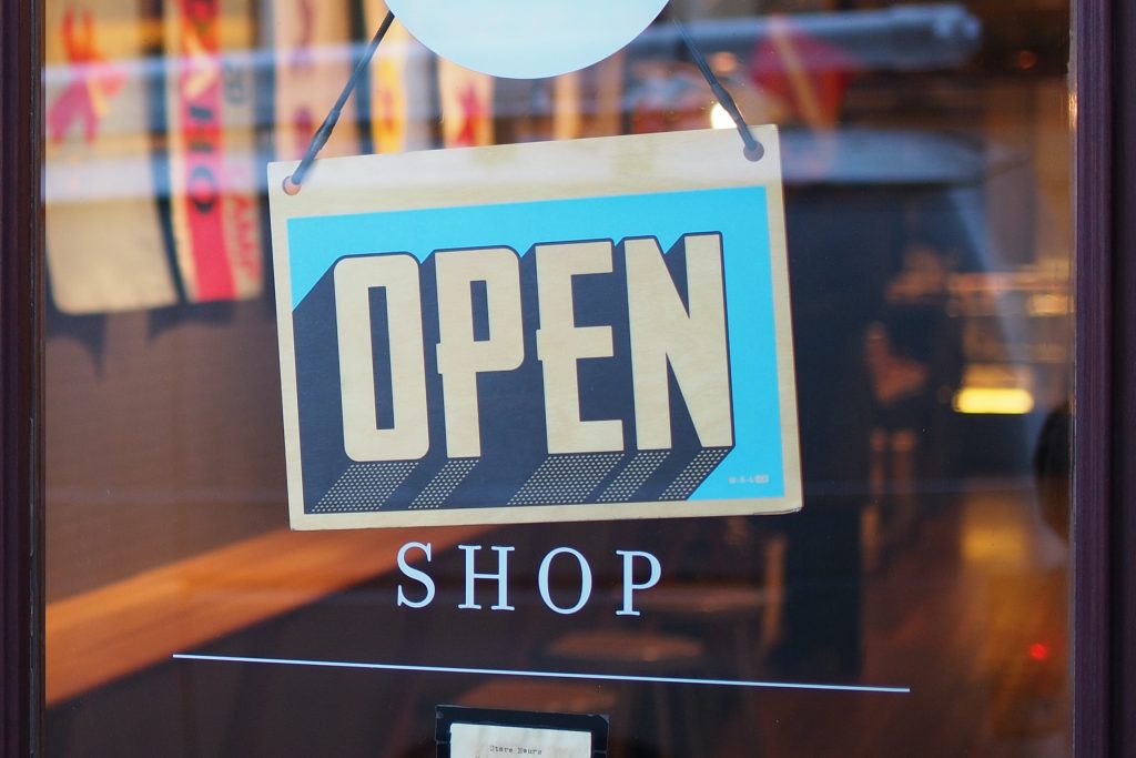 Ein "Geöffnet" Schild hängt an einer Glastür die in einen Kaufladen führt